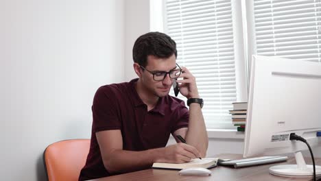 Un-Joven-Empresario-Concentrado-Con-Gafas-Escribiendo-Notas-Mientras-Habla-Por-Teléfono-Móvil-Con-Un-Cliente-En-La-Oficina.-Filmado-En-4k