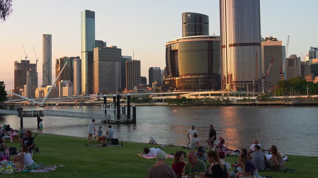 Family and friends gather for a picnic on River Quay Green, South Bank on the weekend, overlooking at Brisbane river views and downtown cityscape at sunset, time-lapse shot.