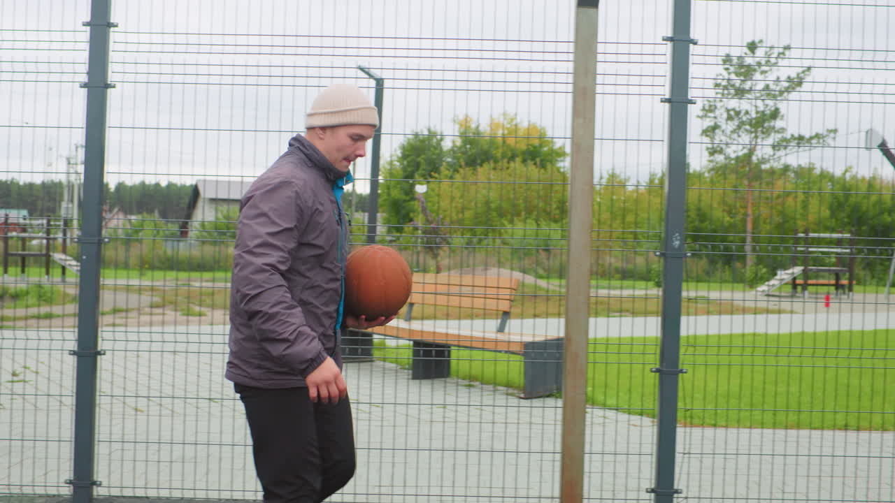 solo man dribbling basketball on outdoor court, wearing beanie and jacket under cloudy sky, bouncing ball toward fenced hoop, moving through warmup and shot practice with focused expression