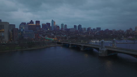 Massive-bridge-over-Charles-river-and-cityscape-at-dusk-on-cloudy-day.-Backwards-descending-fly-above-water-surface.-Boston,-USA