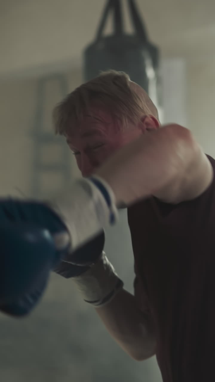 스포츠 센터에서 복싱 가방을 때리는 운동선수, 장갑을 입은 기쁜 운동선수는 연기가 은 체육관에서 팔로 매달린 장비를 때립니다. 느린 동작, 전투 공격 스타일