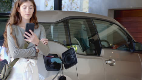 charging electric car, woman using smartphone while waiting at home garage
