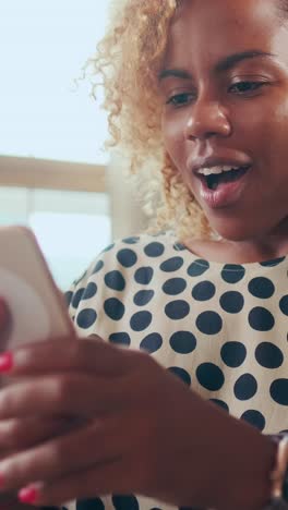 african american woman holding phone and victoriously waving hand sits on sofa