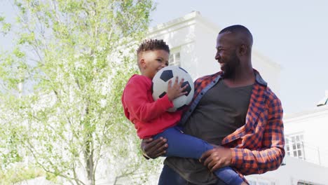Porträt-Eines-Glücklichen-Afroamerikanischen-Vaters-Und-Seines-Sohnes,-Die-Fußball-Halten-Und-Sich-Im-Garten-Umarmen