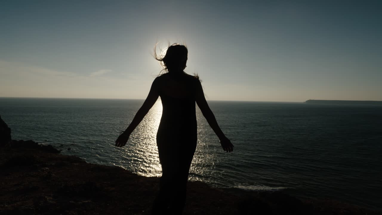 Backlit silhouette of a woman standing on a cliffside in Algarve, Portugal, with sunburst through her hair