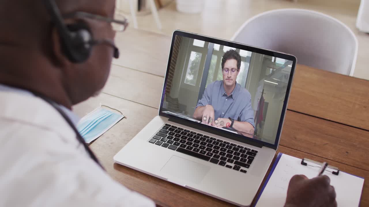 African american male doctor wearing phone headset taking notes while having a video call on laptop