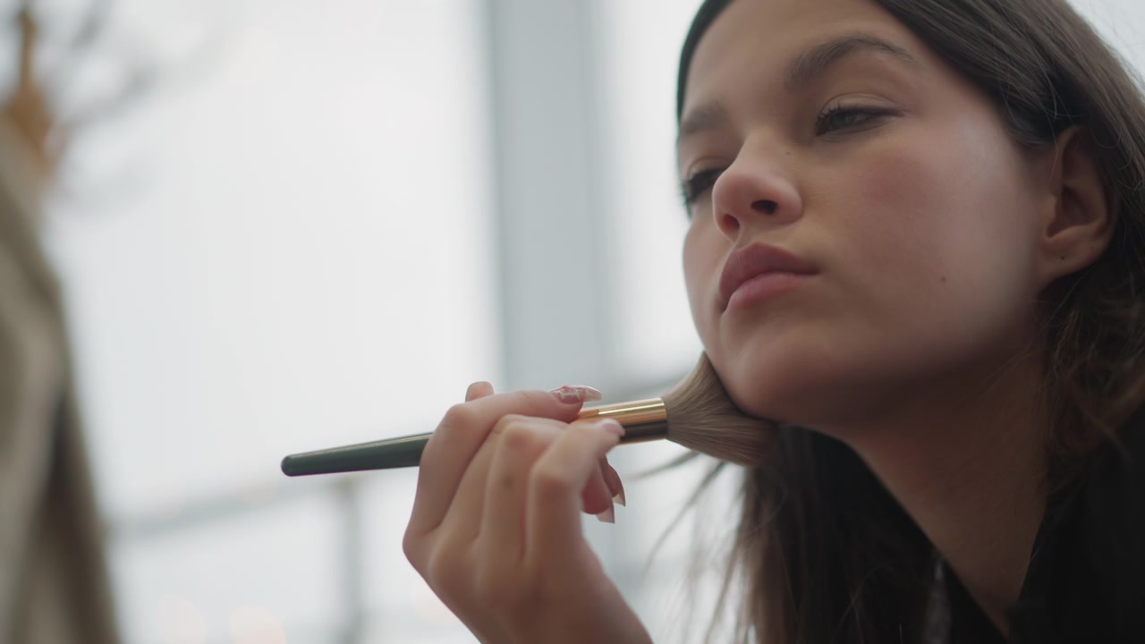 Young woman in bright indoor studio sweeps large makeup brush across cheek, chin, and jawline while tilting head in profile; hand moves in repeated gentle strokes for calm beauty preparation