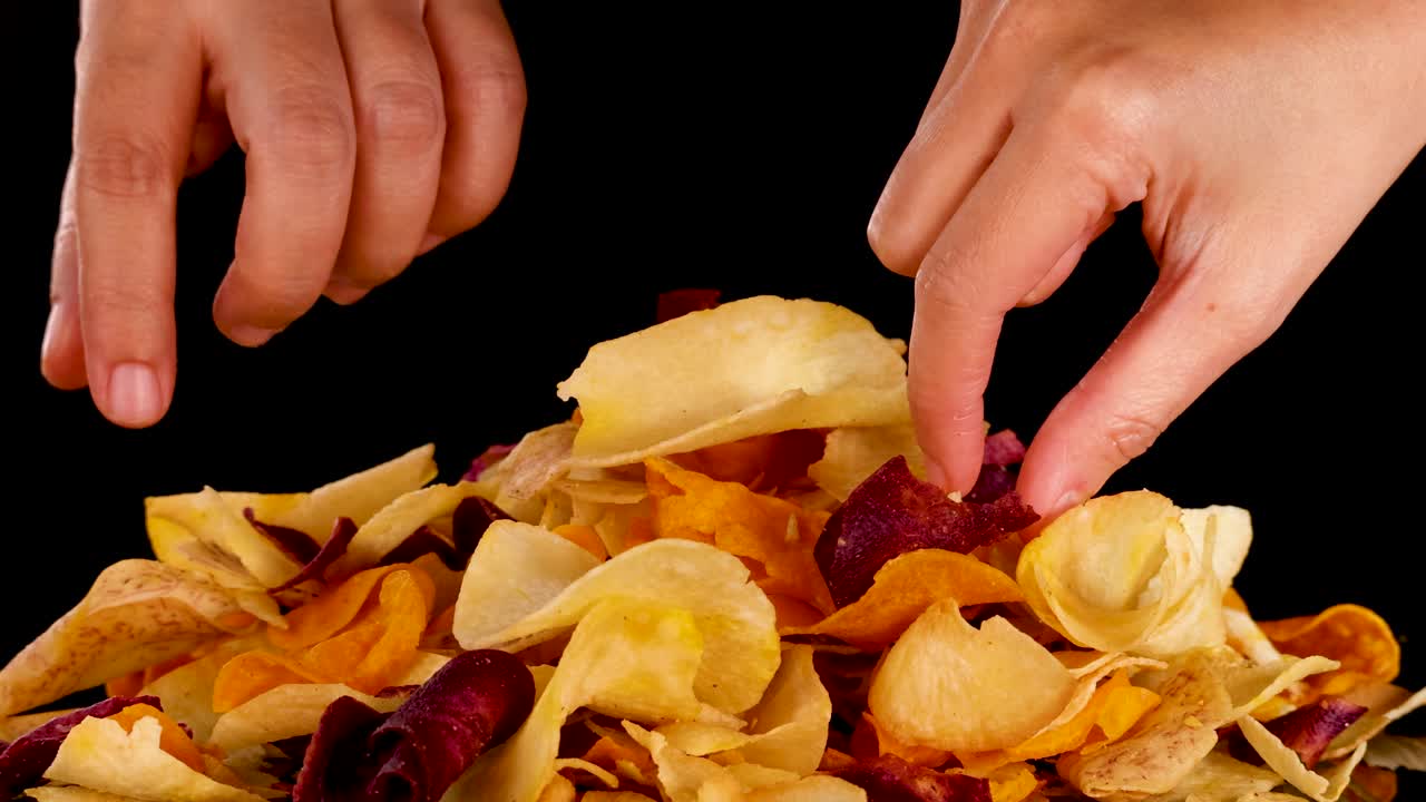 Hand selects crispy mixed root vegetable chips from pile, studio lighting, close-up, black background