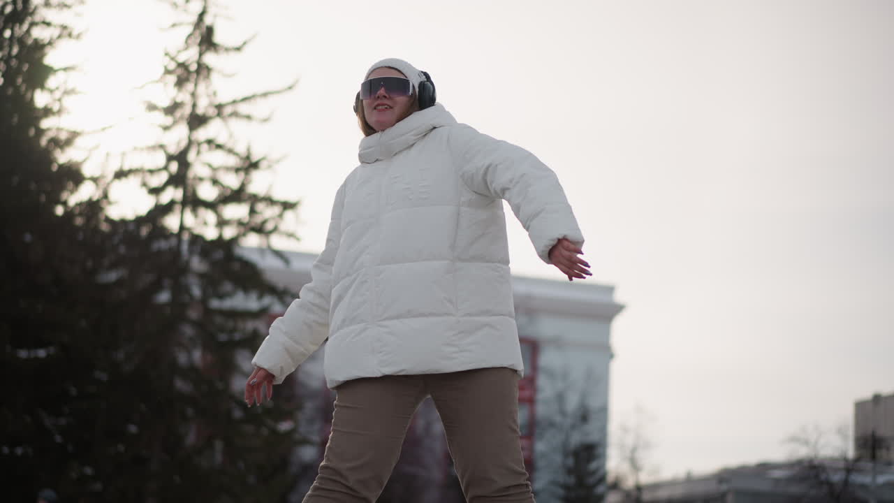 Closeup of teenager wearing white puffer jacket, beanie, black goggles and headphones practicing dance moves with fluid hand and body gestures on snow covered urban park plaza