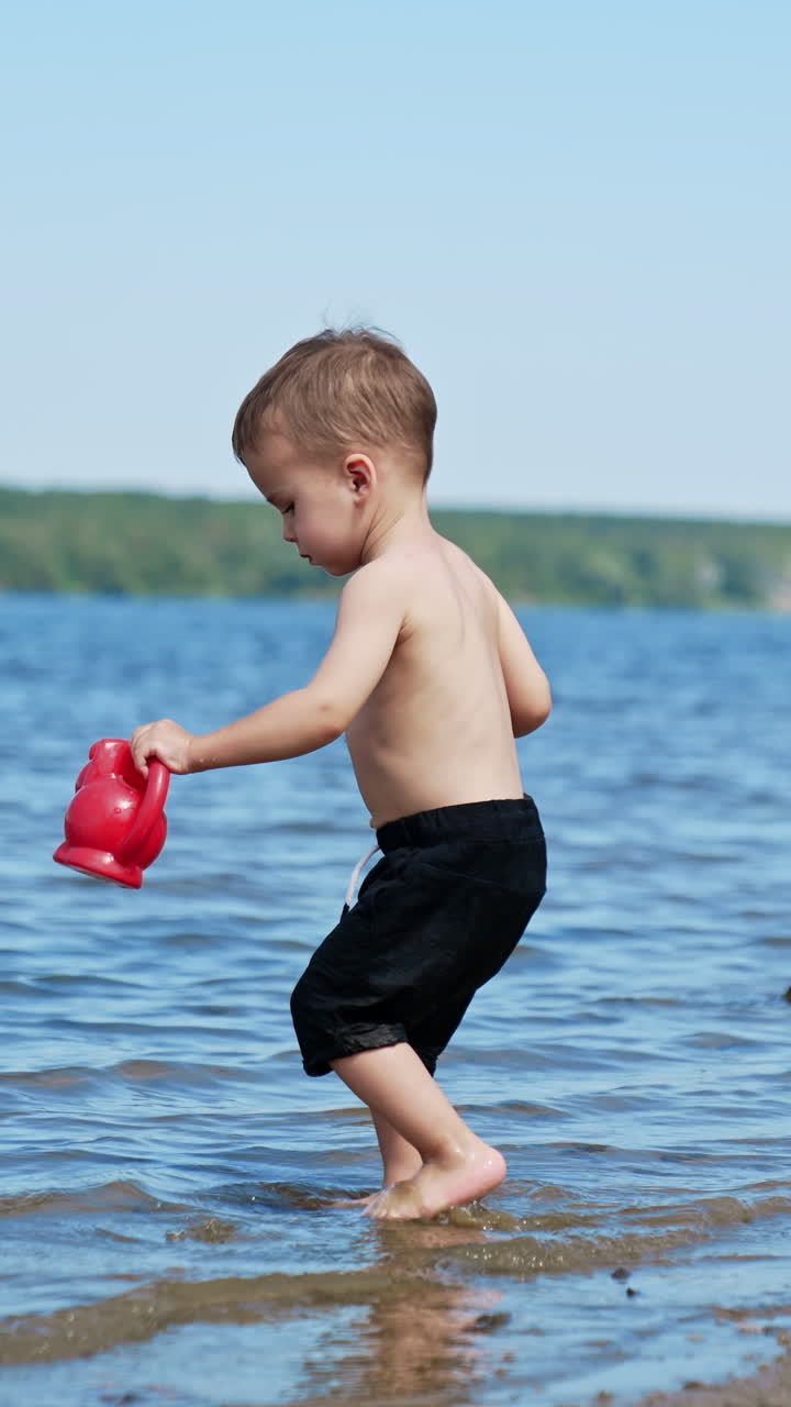 Lovely baby boy spending time at the beach in summer. Toddler walks into the water to fill his watering can. Vertical video