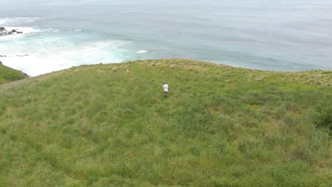 órbita aérea alrededor de un hombre que camina en la hierba verde al borde de un gran acantilado con el gran océano del sur a la vista