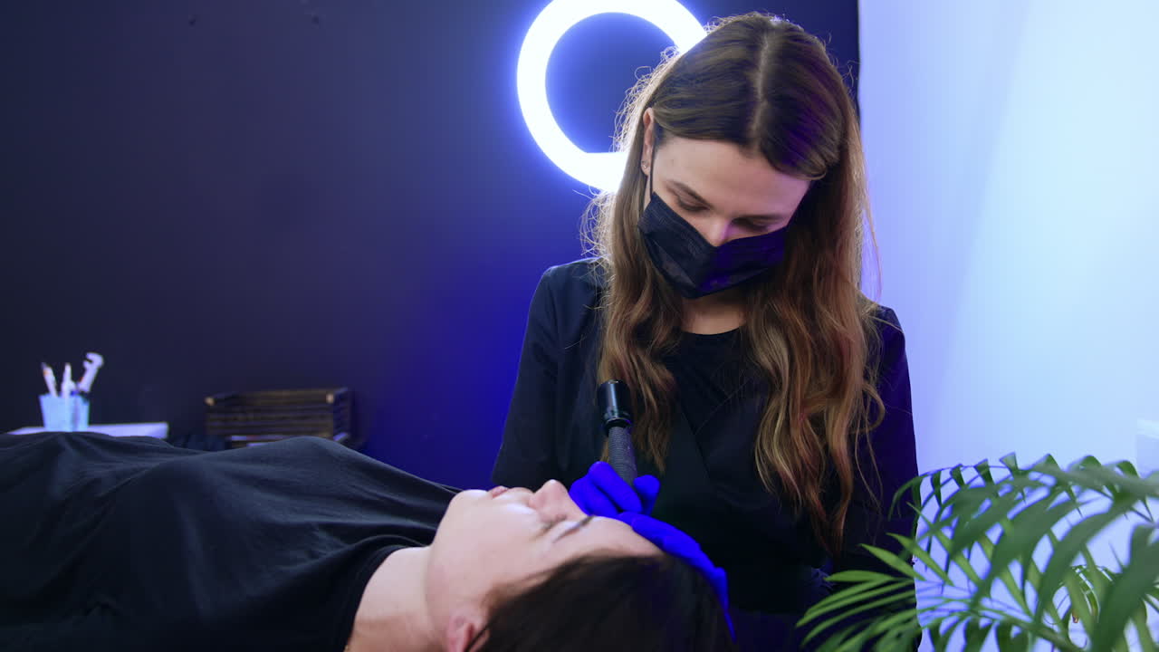 Long-haired brow-master in mask performs the procedure for the client. Beauty salon eyebrow make up for a female client.