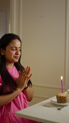 A-happy-brunette-girl-in-a-pink-dress-sits-in-front-of-a-table-in-front-of-a-small-cake-and-blows-out-a-pink-candle-on-it-applauding-and-clapping-her-hands-in-a-modern-apartment