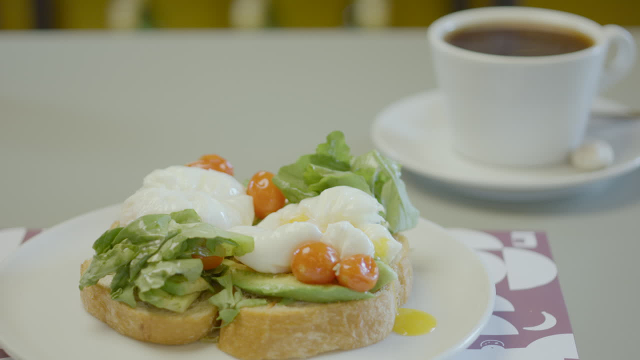 Close-up of a plate with rustic bread toast topped with avocado, lettuce, cherry tomatoes and poached eggs, next to a cup of hot coffee