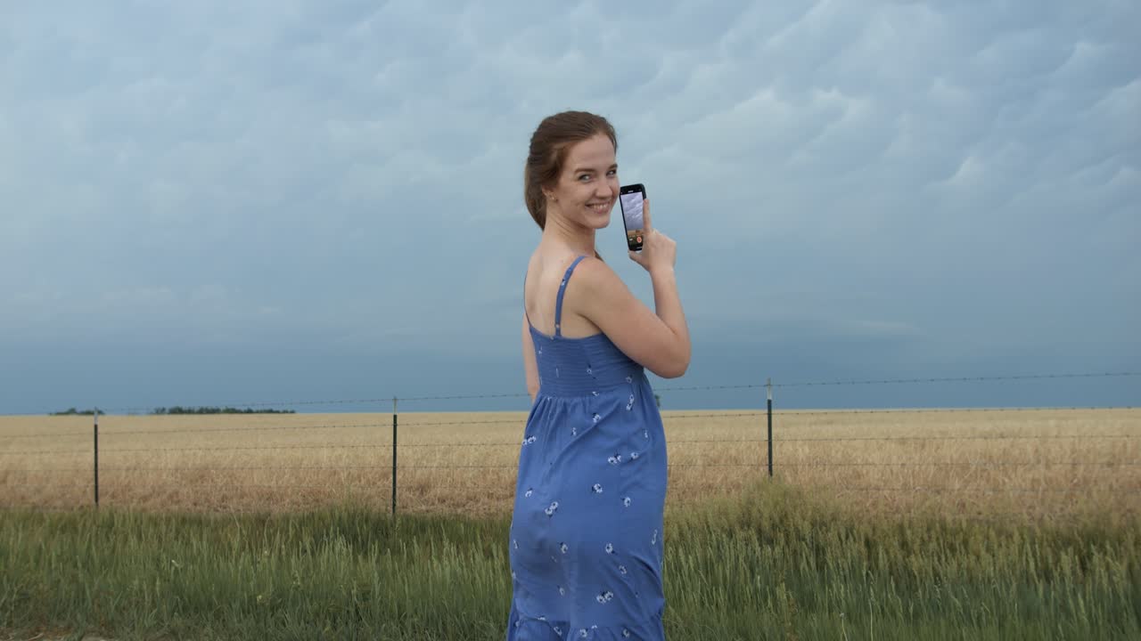 A young woman wearing a blue dress on farm land watching distant storm clouds.