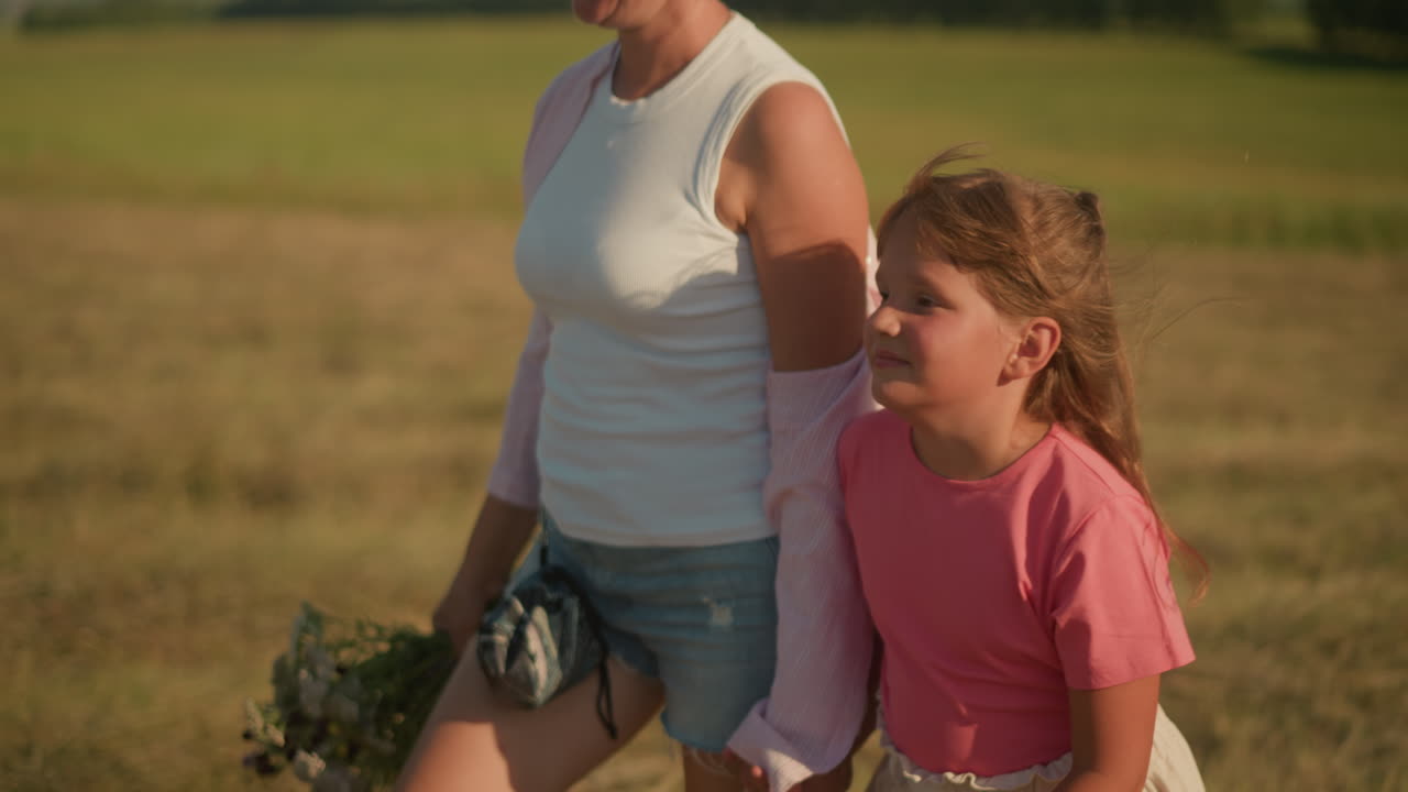 Side view of little girl and aunt walking hand in hand in sunny countryside, aunt holding bouquet of flowers, both smiling warmly, enjoying summer day in open field with blurred green landscape