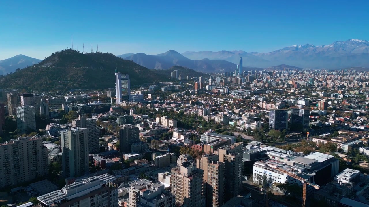 Sorvolo ad alta quota sopra quartieri residenziali che rivela l'intero skyline di Santiago con il Cerro San Cristóbal, il Costanera Center e la catena delle Ande innevate. Santiago, Cile.