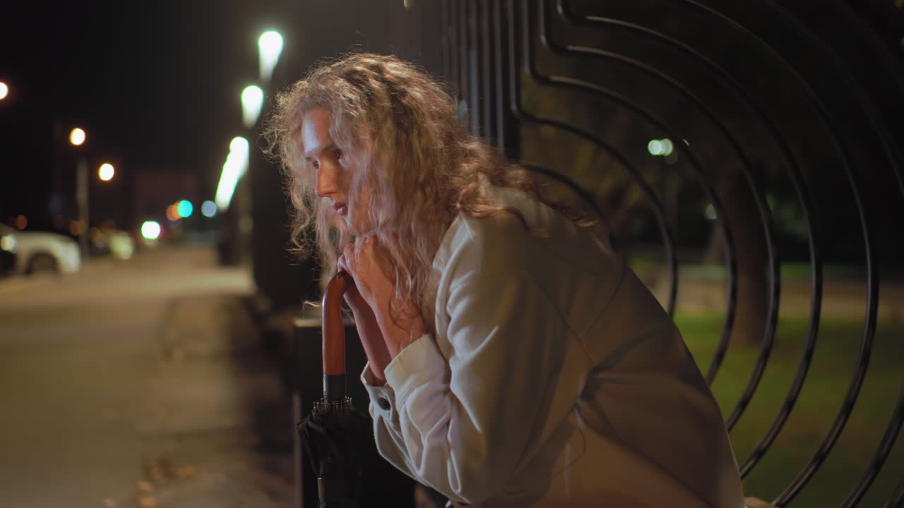 Young girl with curly hair wearing white coat sits alone on decorative wooden bench by iron fence at night, resting on umbrella, calmly waiting for friend with soft glowing streetlights in background