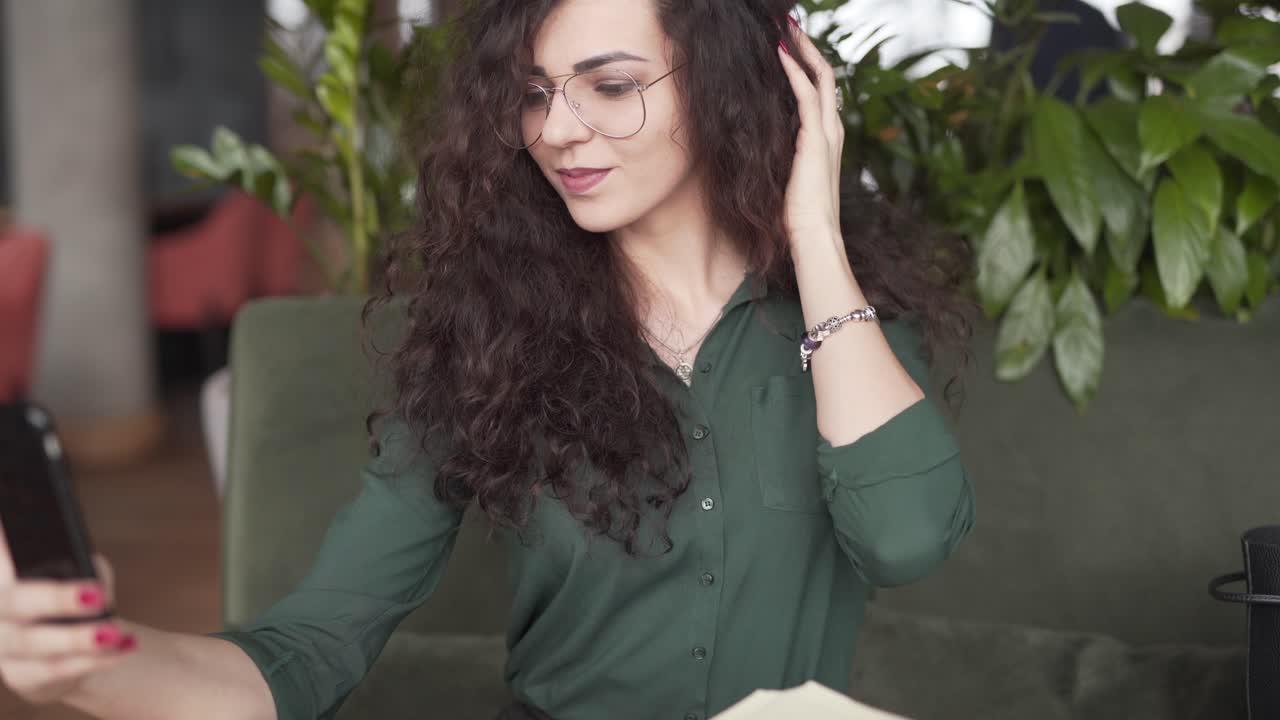 hermosa joven juega con su cabello rizado y se toma una selfie mientras está sentada dentro de un café