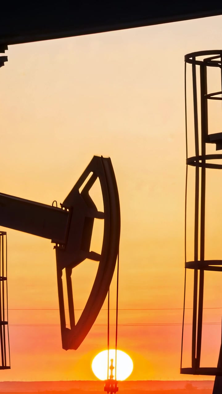 Vertical video: Pumping pumpjack beam lifting horsehead at oilfield, dawn moving rod past sun, tank