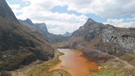 a stunning aerial shot reveals a picturesque pathway winding through a remote mountain valley alongside a rusty brown lake on the epic huayhuash trek in the peruvian andes