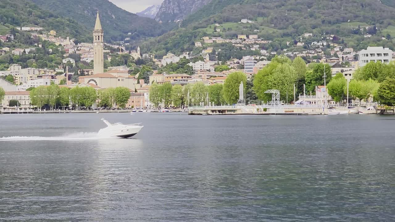 A speedboat is going past the camera on Lake Como. There is a city and mountains in the background.