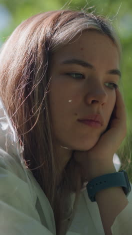 primer plano de una mujer joven con un impermeable transparente, descansando el mentón en la mano en una postura pacífica y contemplativa, con un fondo natural verde suavemente borroso