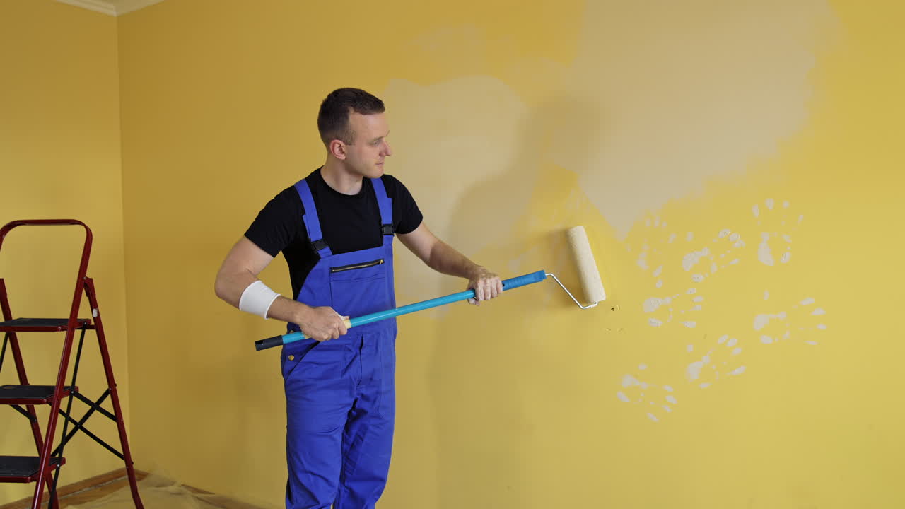 Man is painting walls at home. Young builder in blue uniform renovating walls with a paint roller on a long handle. Painting services.