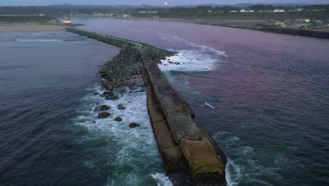 lighthouse in rocky jetty with waves splashing and rising moon during blue hour at background in bandon, oregon