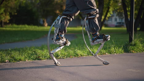 man's legs using spring stilts while walking on paved path in sunny park, wearing gray cargo pants. camera at ground level focuses on stilts and legs, with green grass and trees