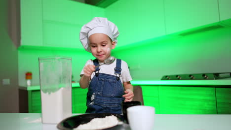 focused caucasian toddler in chef cap uses a spoon to take some flour from a container. little cook in the kitchen.