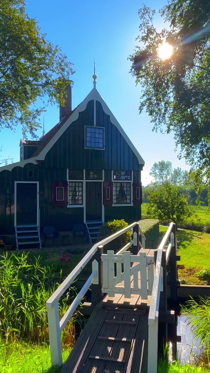 Traditional wooden house with a small bridge surrounded by vibrant green foliage under bright clear blue sky with lens flare in Zaanse Schans, Zaandam, Netherlands.