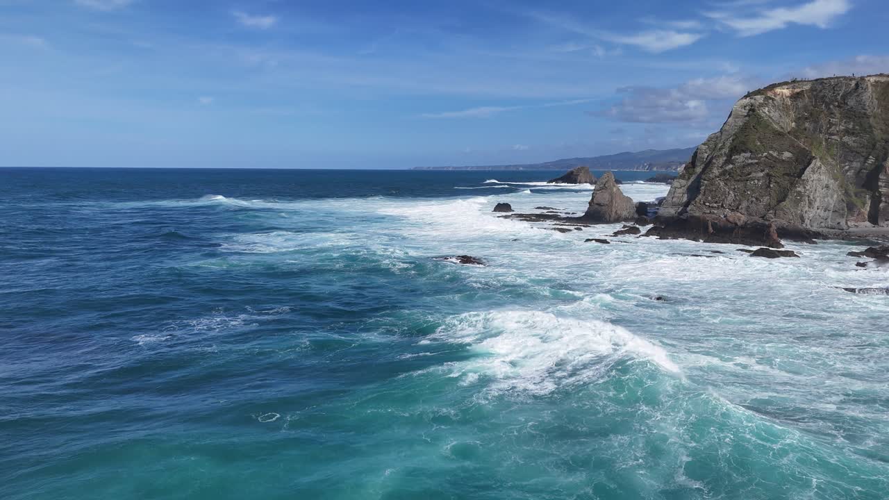 Pemandangan drone air laut pirus, tebing berbatu, dan ombak bergulung di sepanjang garis pantai yang terjal.