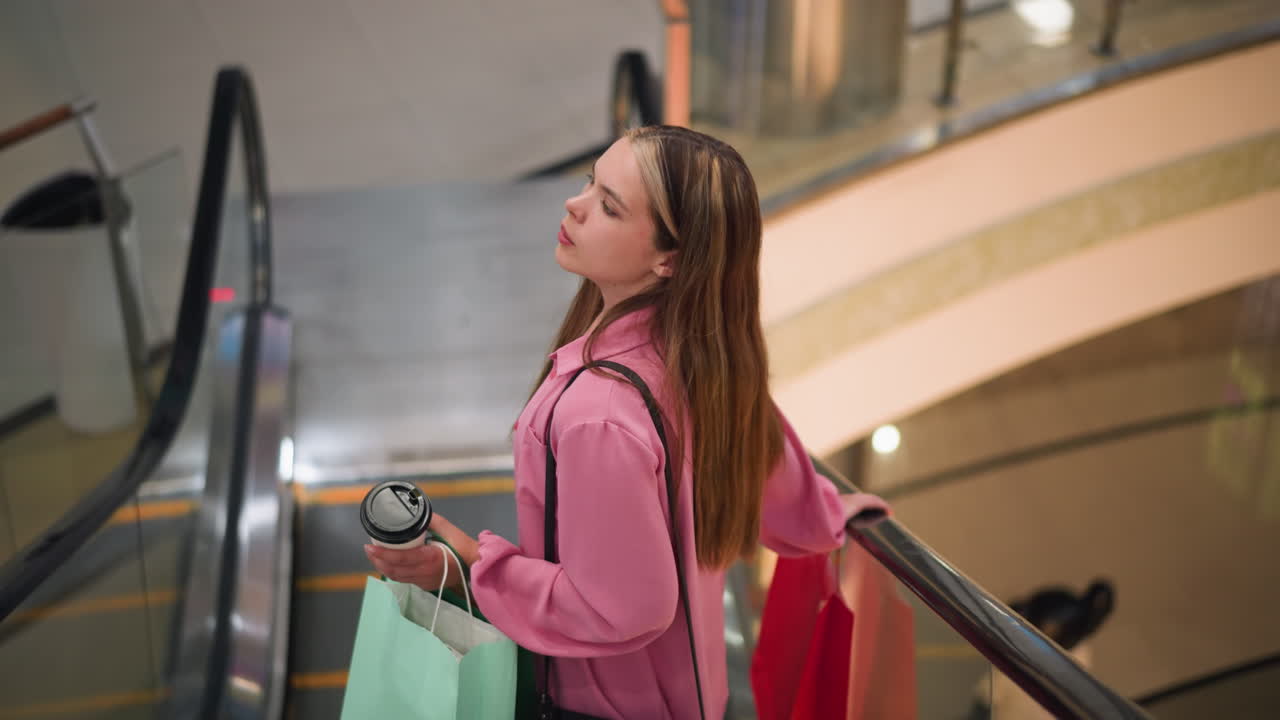 Woman in pink dress holding coffee cup and shopping bags while standing on an escalator as it descends in a brightly lit mall, she appears tired, with colorful wall designs and glass railings visible