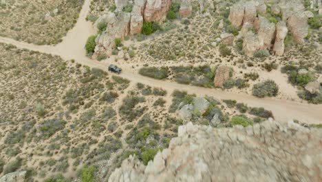 un avión no tripulado vuela alto sobre rocas y sobre el paisaje desértico en el área salvaje de cederberg, sudáfrica, y usted ve un vehículo todoterreno negro desde la vista de un pájaro.