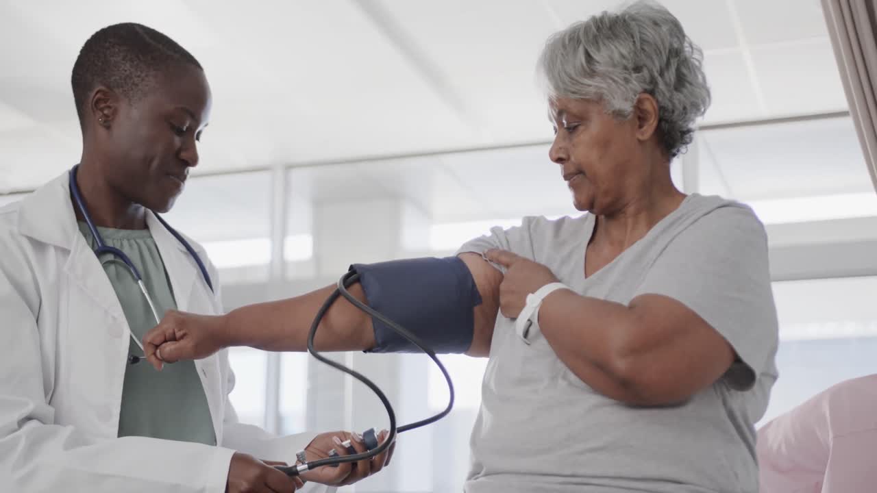 Happy diverse female doctor taking blood pressure of senior female patient in hospital, slow motion