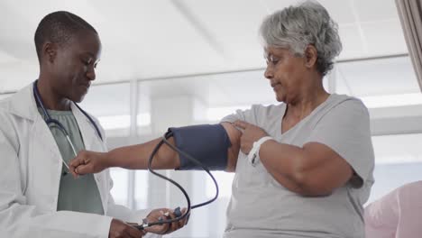 happy diverse female doctor taking blood pressure of senior female patient in hospital, slow motion