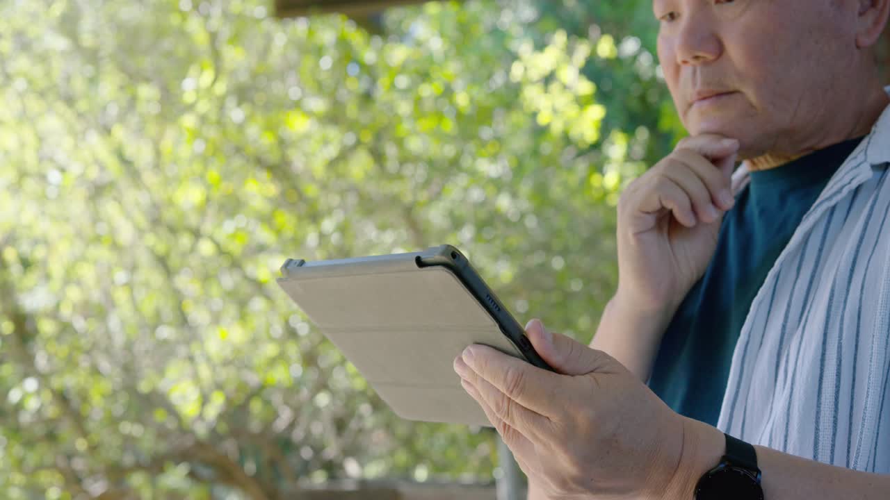Senior Asian man under shelter holding tablet case, tapping, swiping, reading screen, copy space