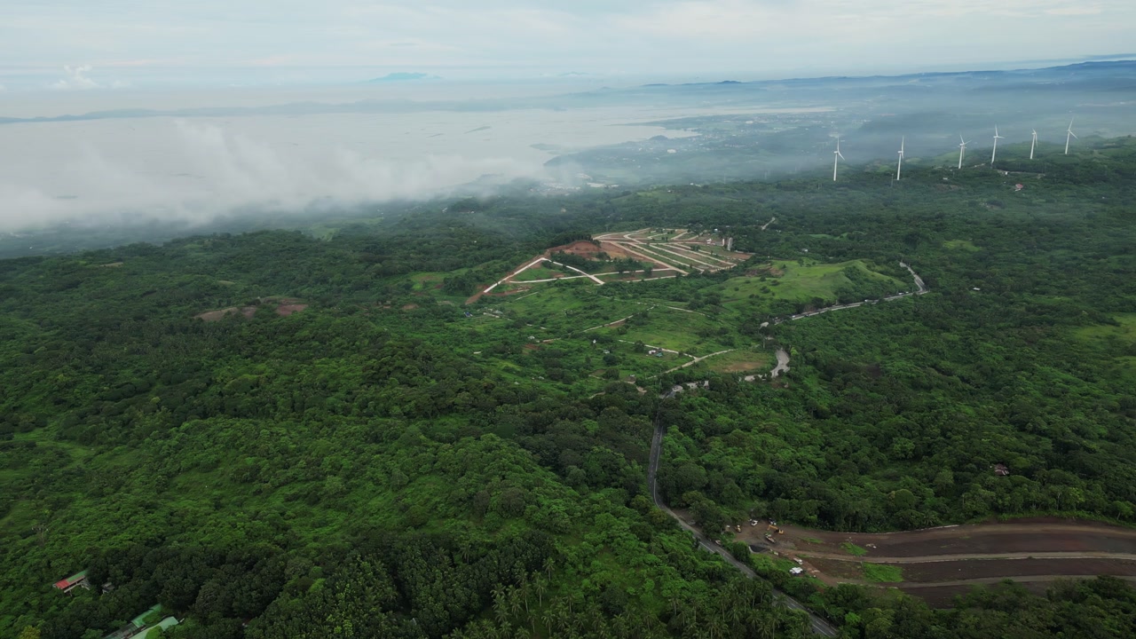 An aerial pan across a lush forested valley, revealing winding roads, cultivated plots, and wind turbines rising through mist in a blend of nature in Mauban, Quezon Province Philippines