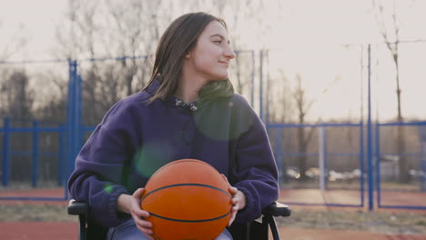 Glückliche-Behinderte-Frau-Im-Rollstuhl,-Die-In-Die-Kamera-Schaut,-Während-Sie-Einen-Basketball-Auf-Dem-Basketballplatz-Hält