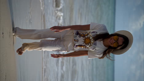 Vertical-slow-motion-of-a-beautiful-latin-brunette-woman-walking-on-the-beach-towards-the-camera-wearing-linen-pants-a-white-hat-and-yellow-sunglasses