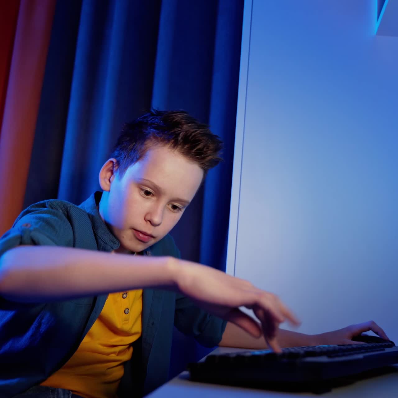 Portrait of teenager playing games. Happy boy sitting in his room and pushing the keyboard buttons while playing on PC. Blue evening light.