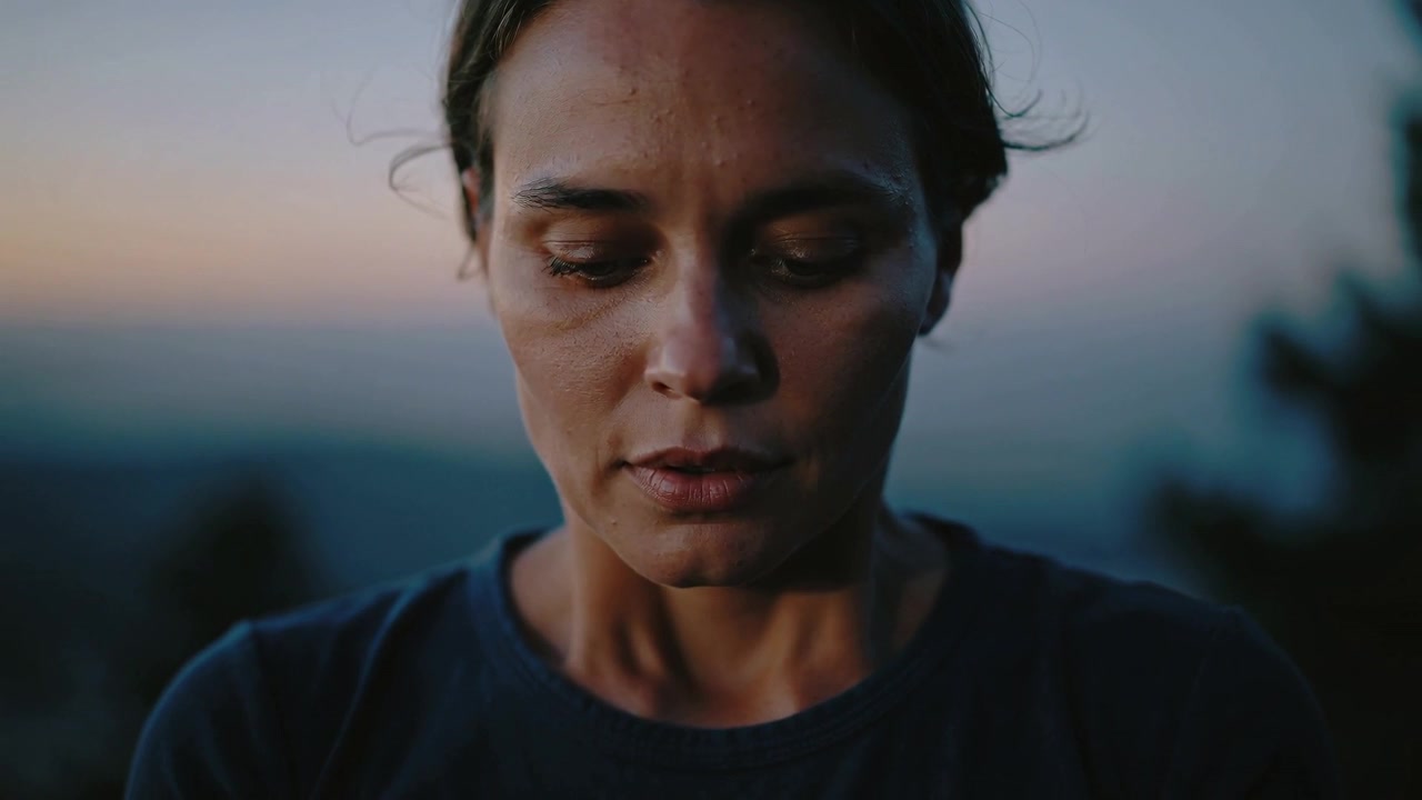 Mulher de blusa escura piscando, abaixando a cabeça e respirando em um mirante ao entardecer, refletindo.