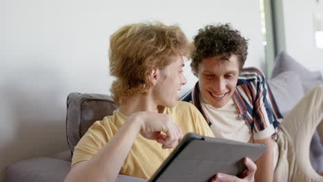 happy diverse gay male couple sitting at sofa using tablet at home, slow motion