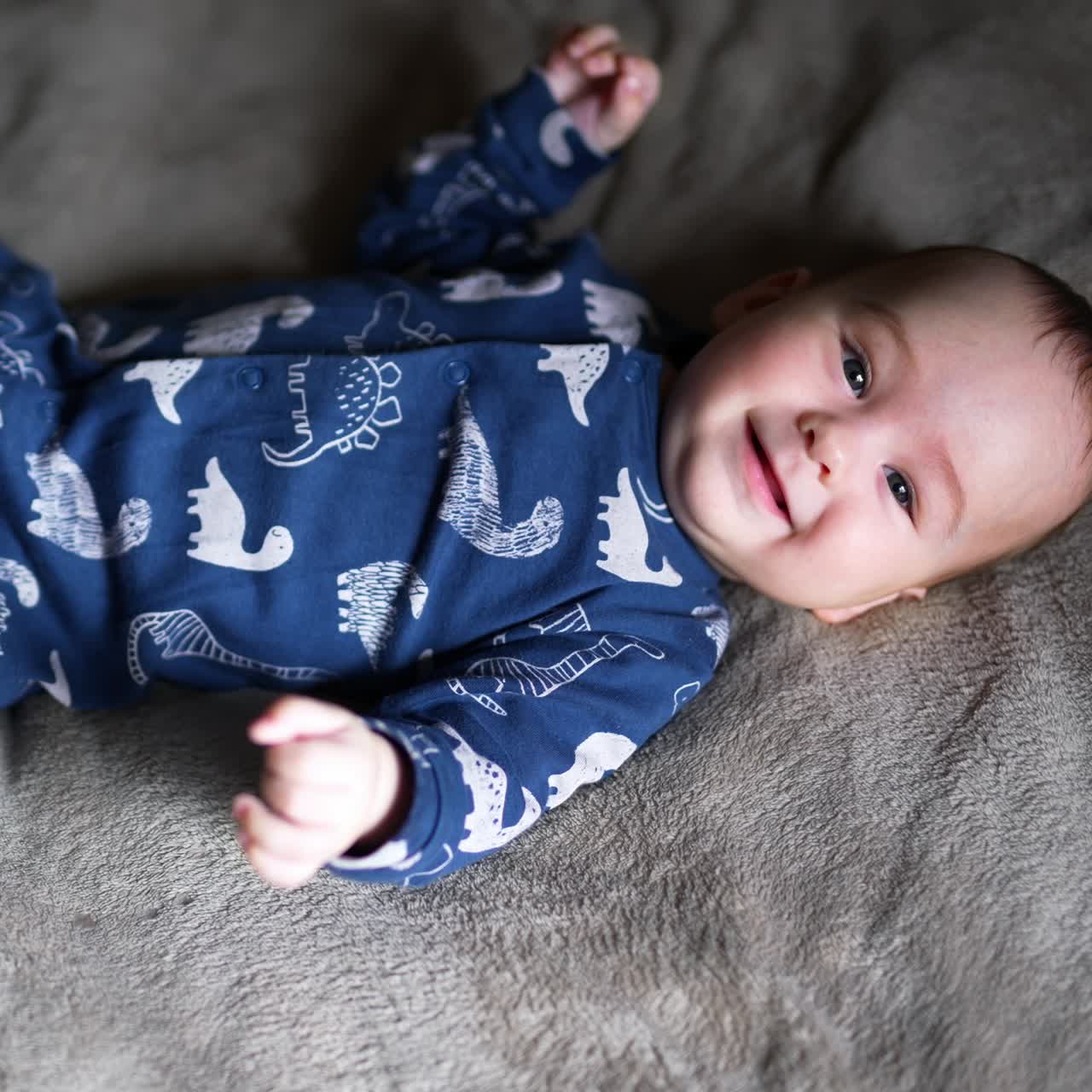 Sweet healthy toddler boy in blue clothes lies on bed. Adorable boy looking into camera and smiling sweetly. Top view