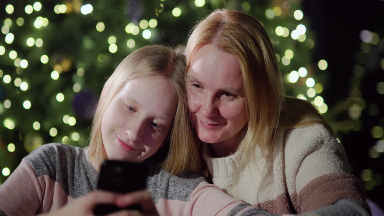 엄마와 딸은 함께 스마트폰 화면을 보고 있습니다. 크리스마스 트리의 흐릿한 불빛 앞에 앉아