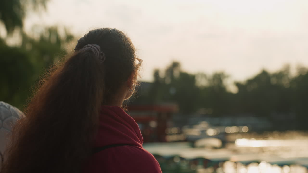 Young asian woman at waterfront dusk, traveler scanning horizon, daydreamer with sketchbook, commuter watching ferries, student pausing to reflect, shimmering water highlights, soft rimlight on hair