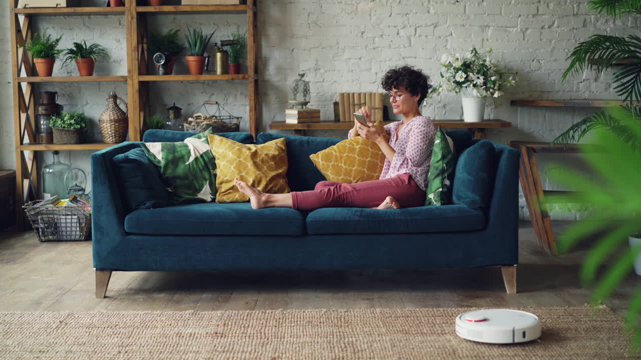 Woman relaxing on a couch using a smartphone in a modern living room with a robotic vacuum cleaner.