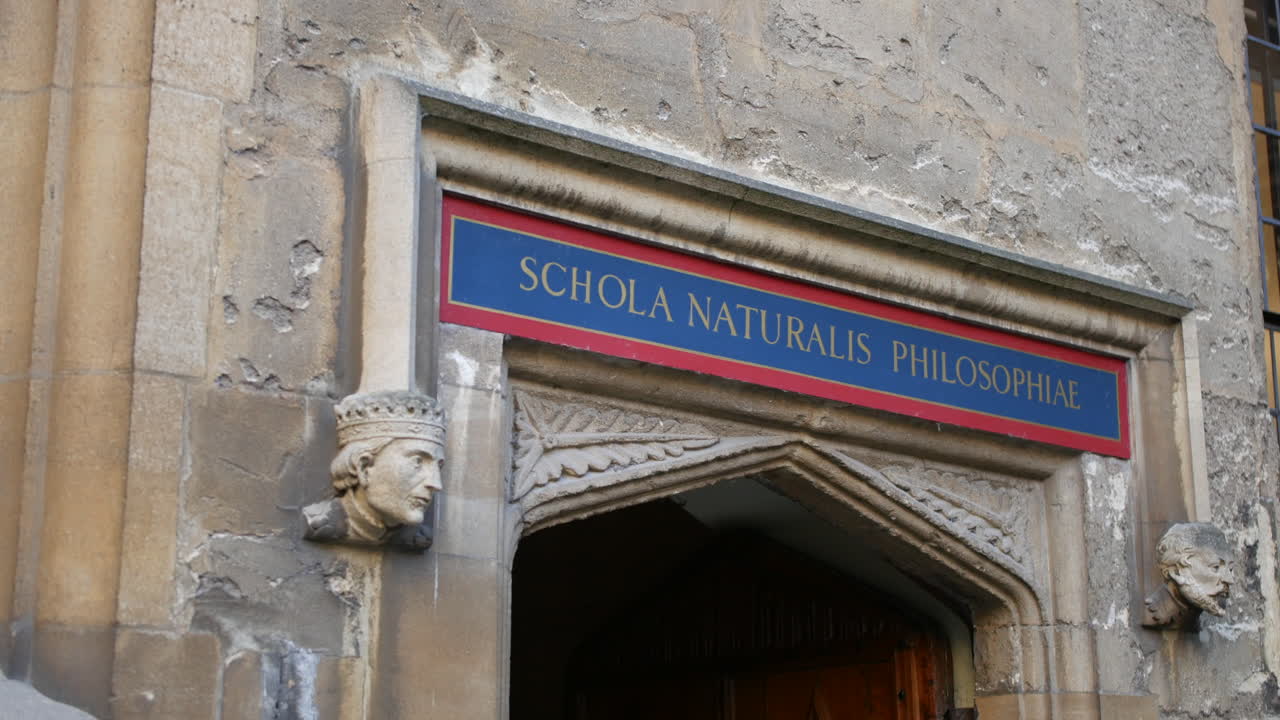 Un antico portale in pietra dell'Università di Oxford con "SCHOLA NATURALIS PHILOSOPHIAE" Scuola di Filosofia Naturale sopra, fiancheggiato da teste scolpite—perfetto per scopi accademici, turismo storico o educativi