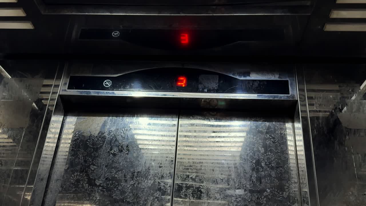 Inside an elevator cabin as doors open to a building hallway, showing stainless steel panels and a red LED floor indicator in soft indoor lighting